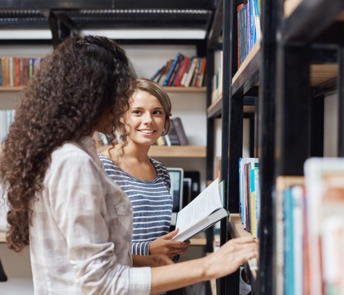 Pair of young beautiful girls in casual stylish clothes standing near bookshelves in library, looking at each other, talking about university life trying to find literature for tomorrow's lesson
