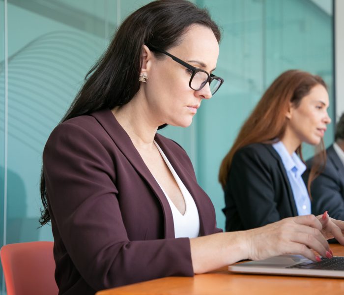 Row of serious business people using laptops, sitting at one table and typing. Woman working at computer near colleagues. Communication or internet technology concept